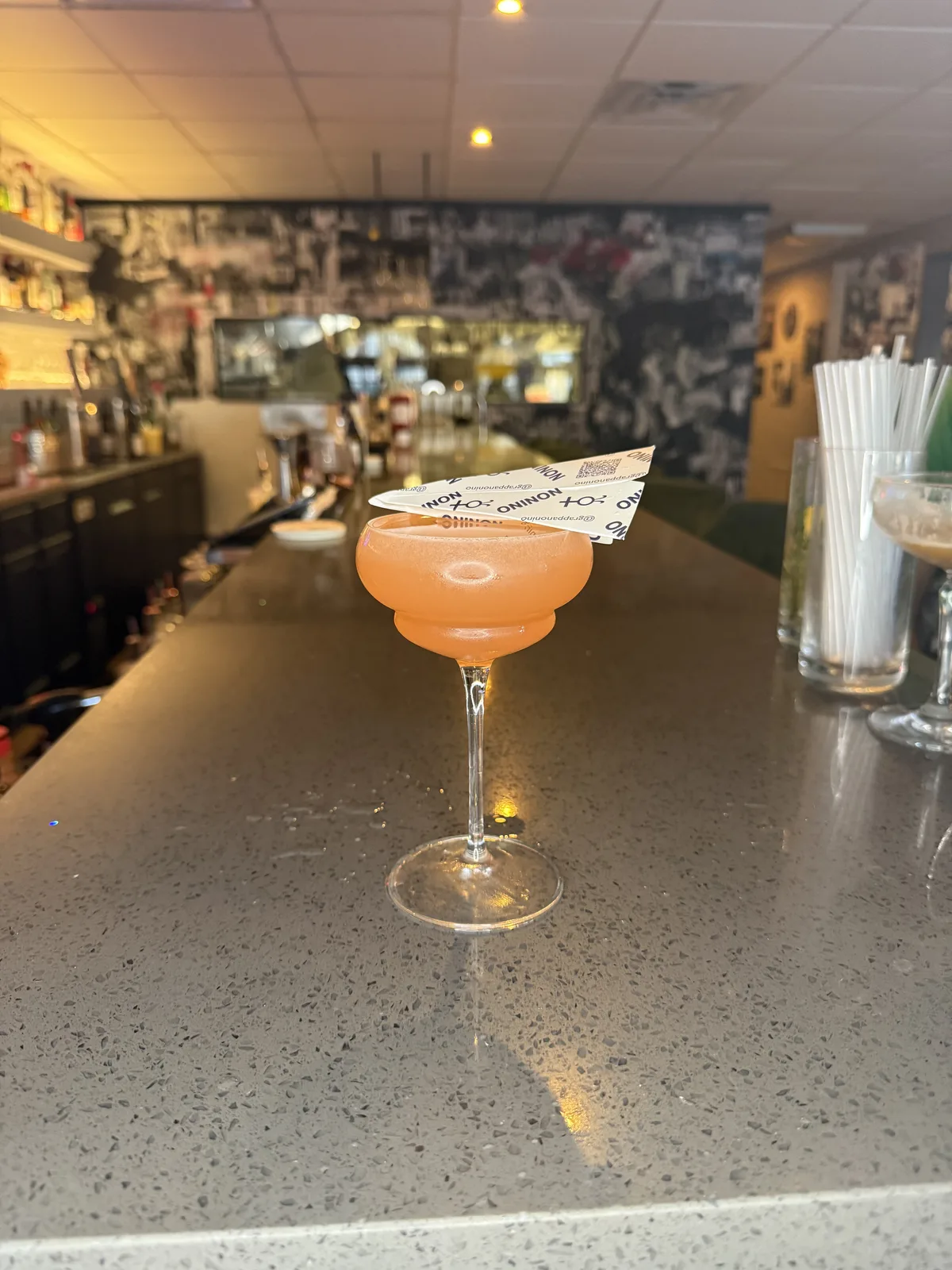 A coupe of pale pink cocktail on the bar counter, the back bar and collage wall behind it