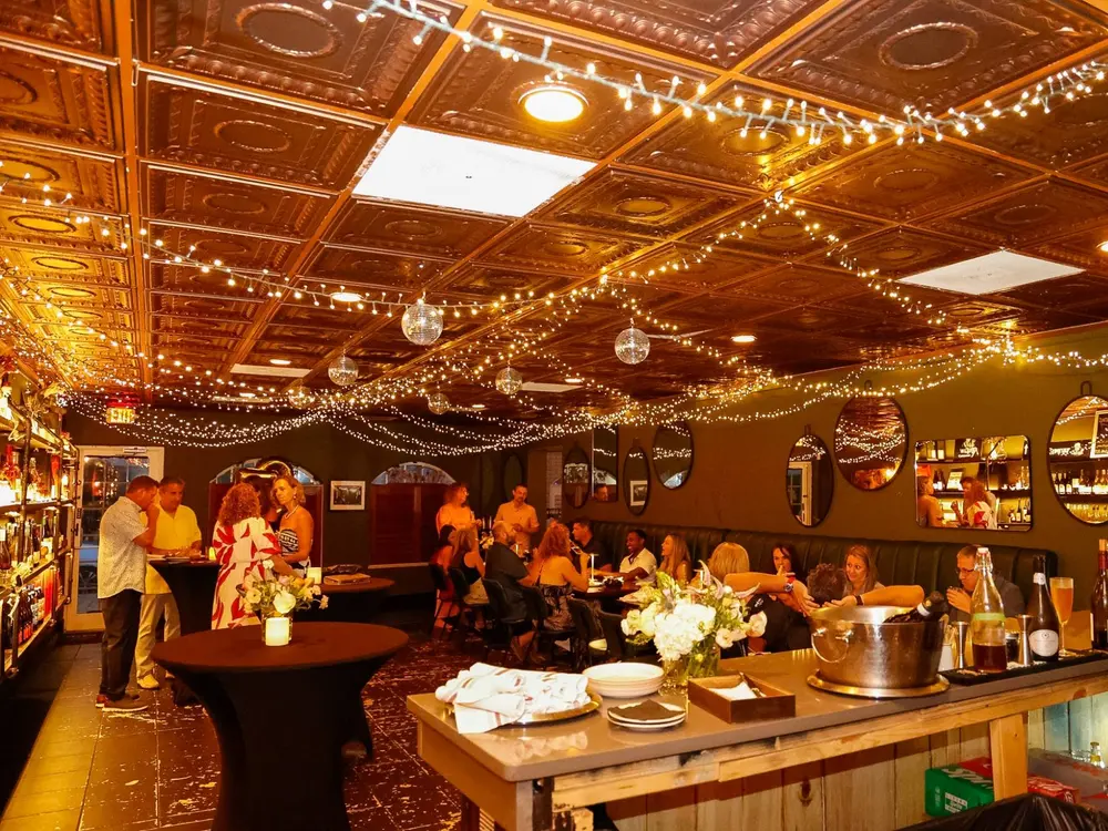 The private room full for a celebration — guests along the banquette and at the bar under string lights and a pressed-tin ceiling